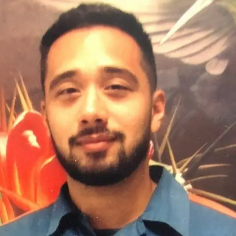 A young man with short dark hair and a beard is wearing a blue collared shirt and standing in front of a colorful, abstract background with red and orange shapes.