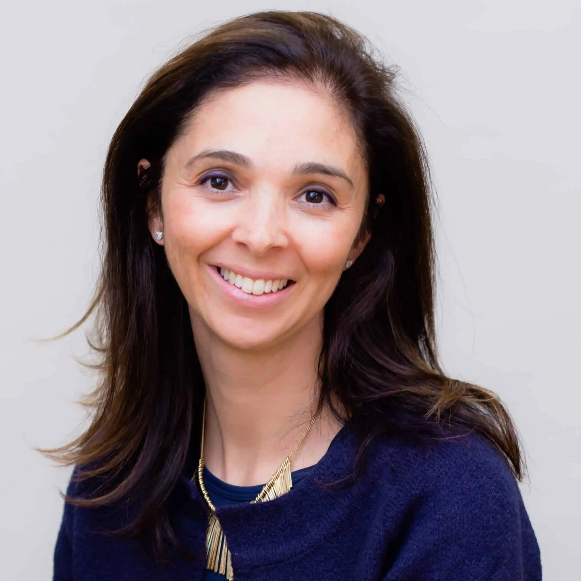 A woman with long brown hair wearing a navy blue top and a gold necklace smiles at the camera, set against a plain light background.
