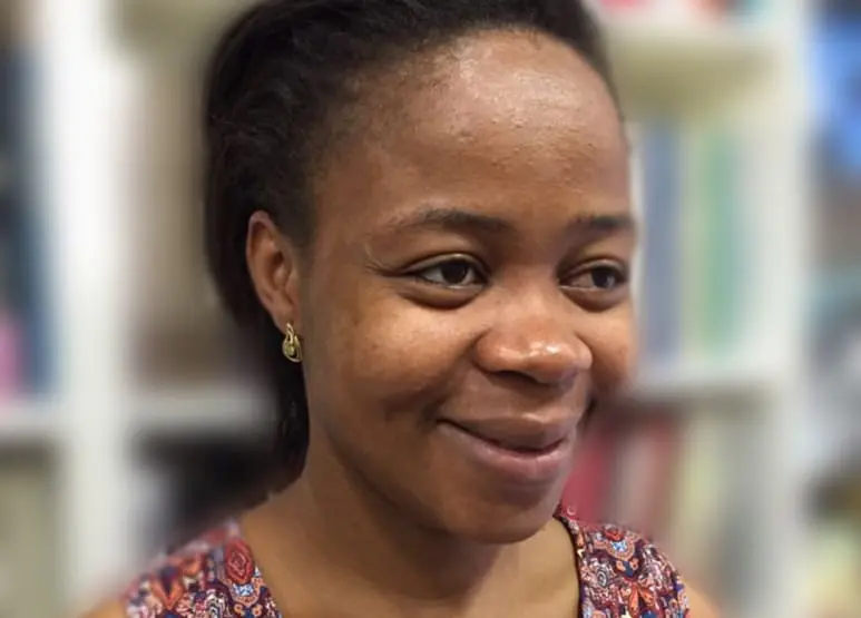 A woman with short dark hair smiles softly. She wears a sleeveless, patterned top and small earrings. The blurred background reveals shelves filled with pride month literature in various colors.