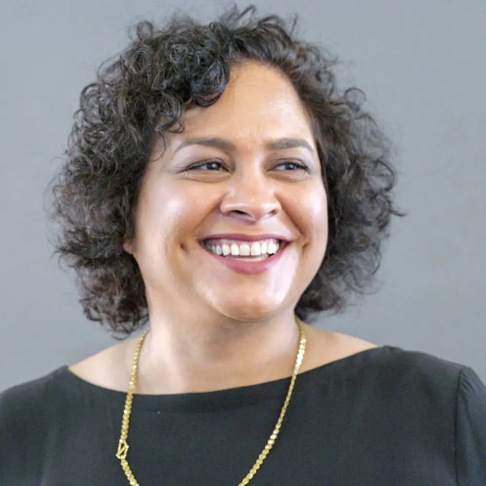 A woman with short curly hair, wearing a black top and a gold necklace, smiles while looking slightly to the side against a plain gray background.