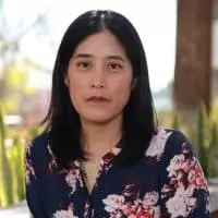 A woman with straight, dark hair wearing a dark floral blouse sits outdoors in front of blurred greenery and sunlight, looking directly at the camera with a neutral expression.