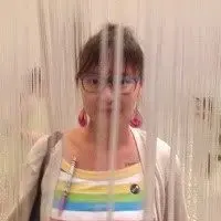 A person wearing glasses and a colorful striped shirt stands behind vertical hanging strings, which partially obscure their face. The background is neutral and out of focus.