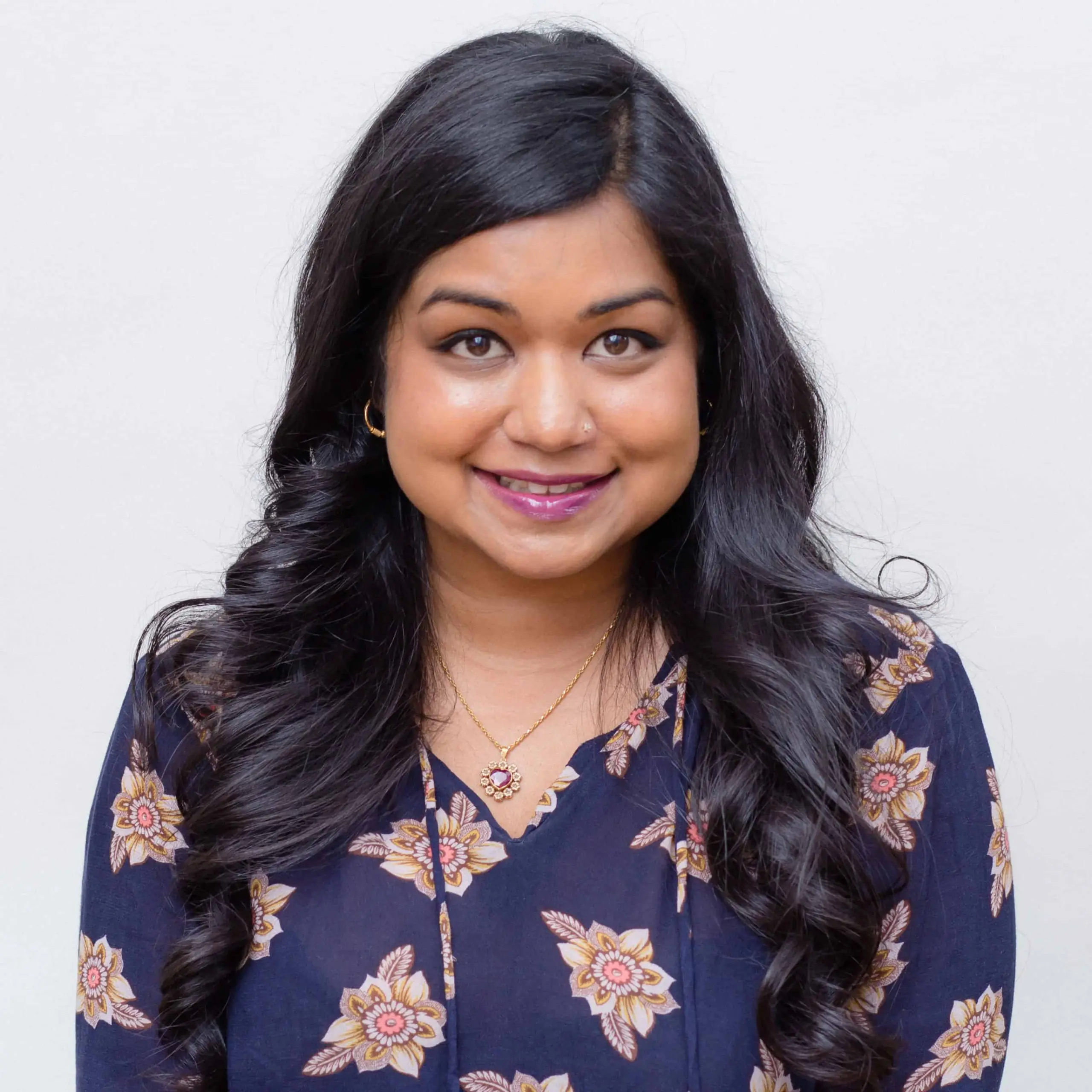 A woman with long, wavy black hair and medium skin tone smiles at the camera. She wears a navy blue floral top, gold hoop earrings, and a gold necklace with a pink pendant, set against a plain light background.
