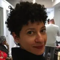 A person with short, curly dark hair smiles slightly while looking over their shoulder in an indoor setting, with other people visible in the background.