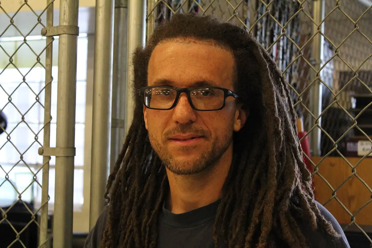 A man with long brown dreadlocks and glasses stands in front of a chain-link fence, wearing a dark shirt. The background is indoors with some light coming through a window.