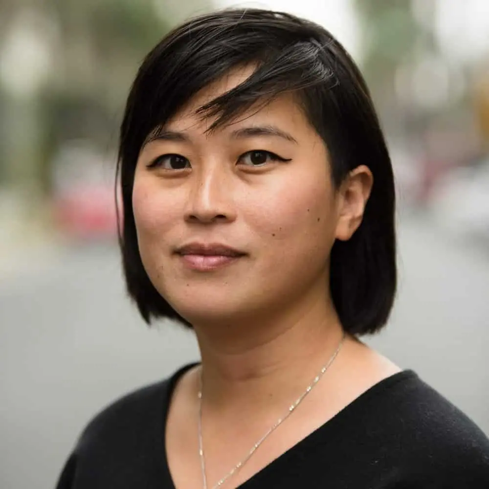 A person with straight, dark hair and side-swept bangs, wearing a black top and a silver necklace, stands outdoors with a blurred background of trees and buildings.