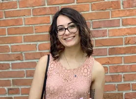 A young woman with wavy brown hair and glasses smiles in front of a brick wall. She is wearing a sleeveless peach lace dress and a necklace, and is holding a small bouquet of flowers.