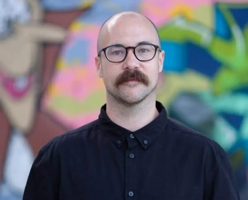 A man with a bald head, glasses, and a thick mustache wearing a black button-up shirt stands in front of a colorful, abstract mural.