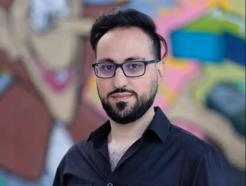 A man with glasses, a beard, and dark hair wearing a black shirt stands in front of a colorful, blurred graffiti mural.