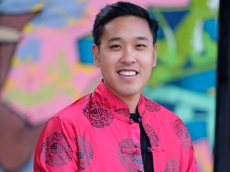 A man smiles at the camera while wearing a bright red traditional jacket with black patterns, standing in front of a colorful, blurred mural background.