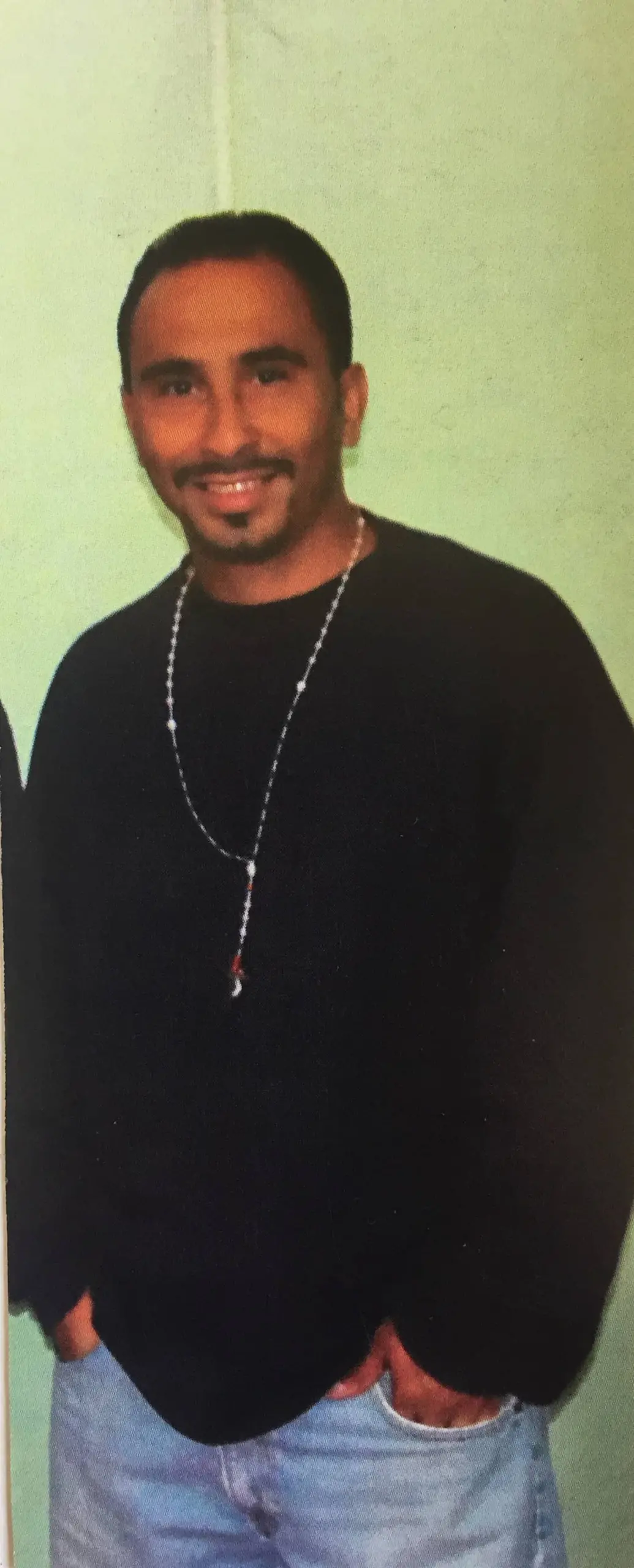 A man with short dark hair and a goatee smiles at the camera. He is wearing a black long-sleeve shirt, a long beaded necklace, and light blue jeans, standing against a light green wall with his hands in his pockets.