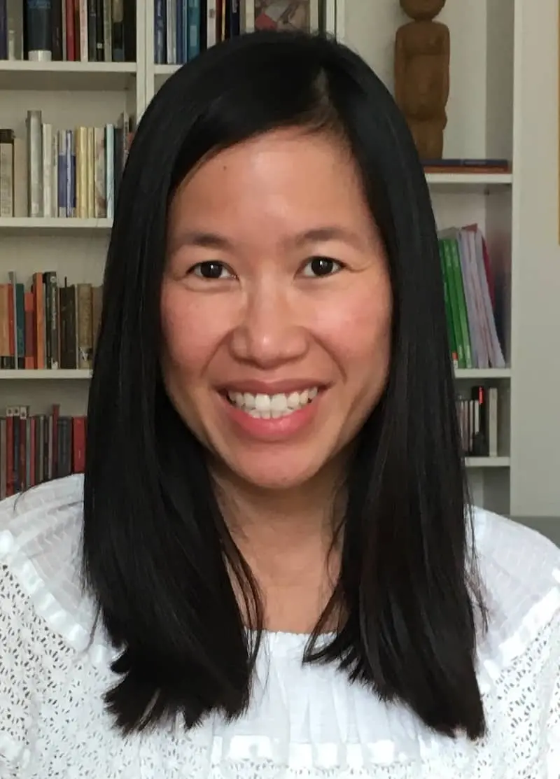 A woman with long straight black hair and a white top smiles at the camera, with a bookshelf filled with books and a wooden sculpture in the background.