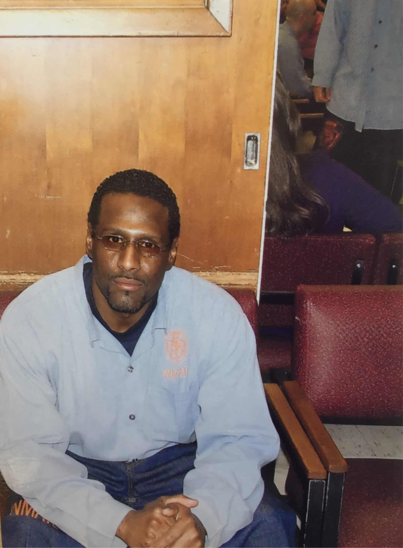 A man wearing glasses and a light blue shirt sits on a chair with his hands clasped. Behind him are maroon chairs, a wooden wall, and a partial reflection in a mirror.