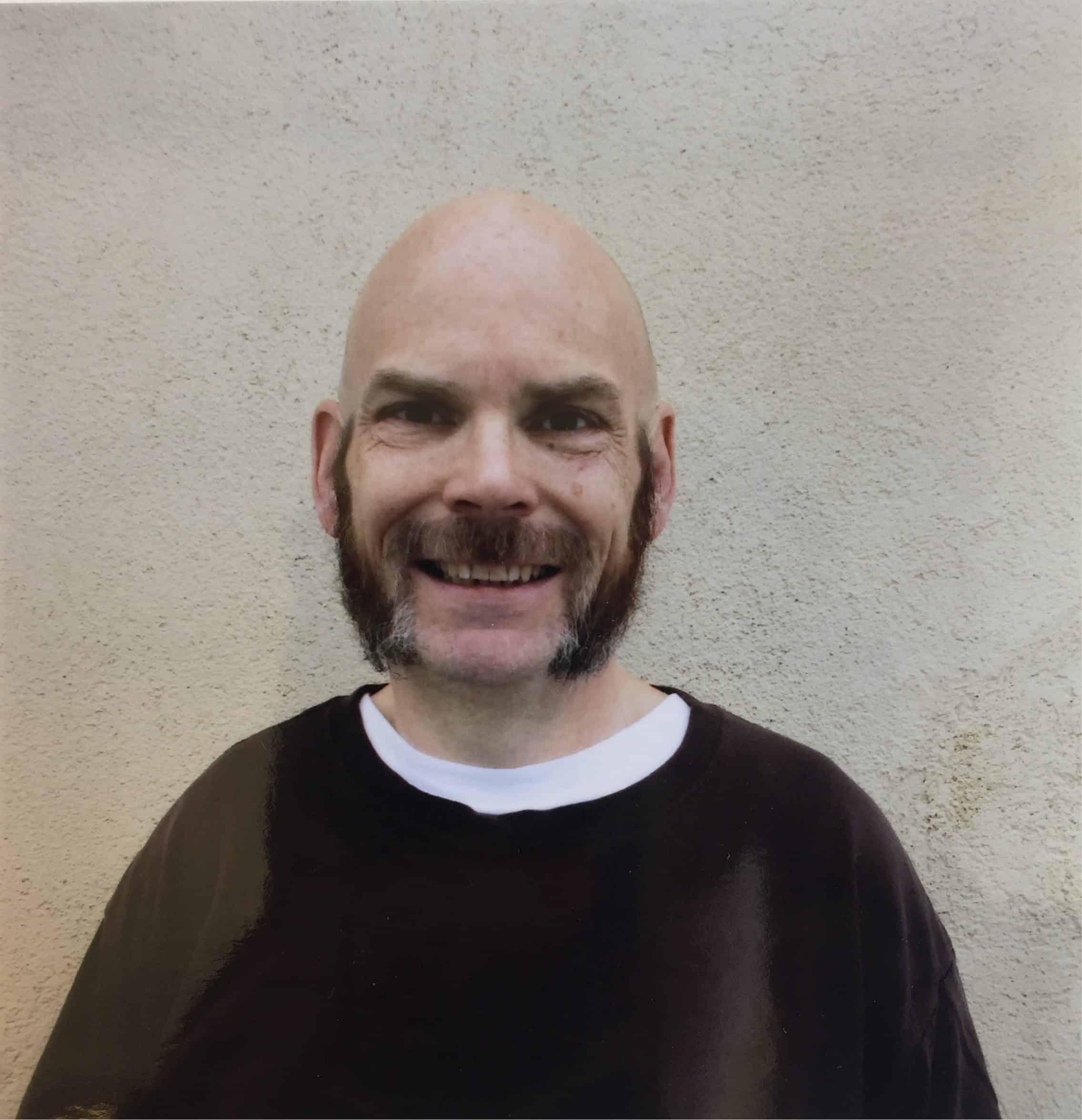 A smiling bald man with mutton chop sideburns and a mustache, wearing a black shirt over a white undershirt, stands in front of a beige textured wall.