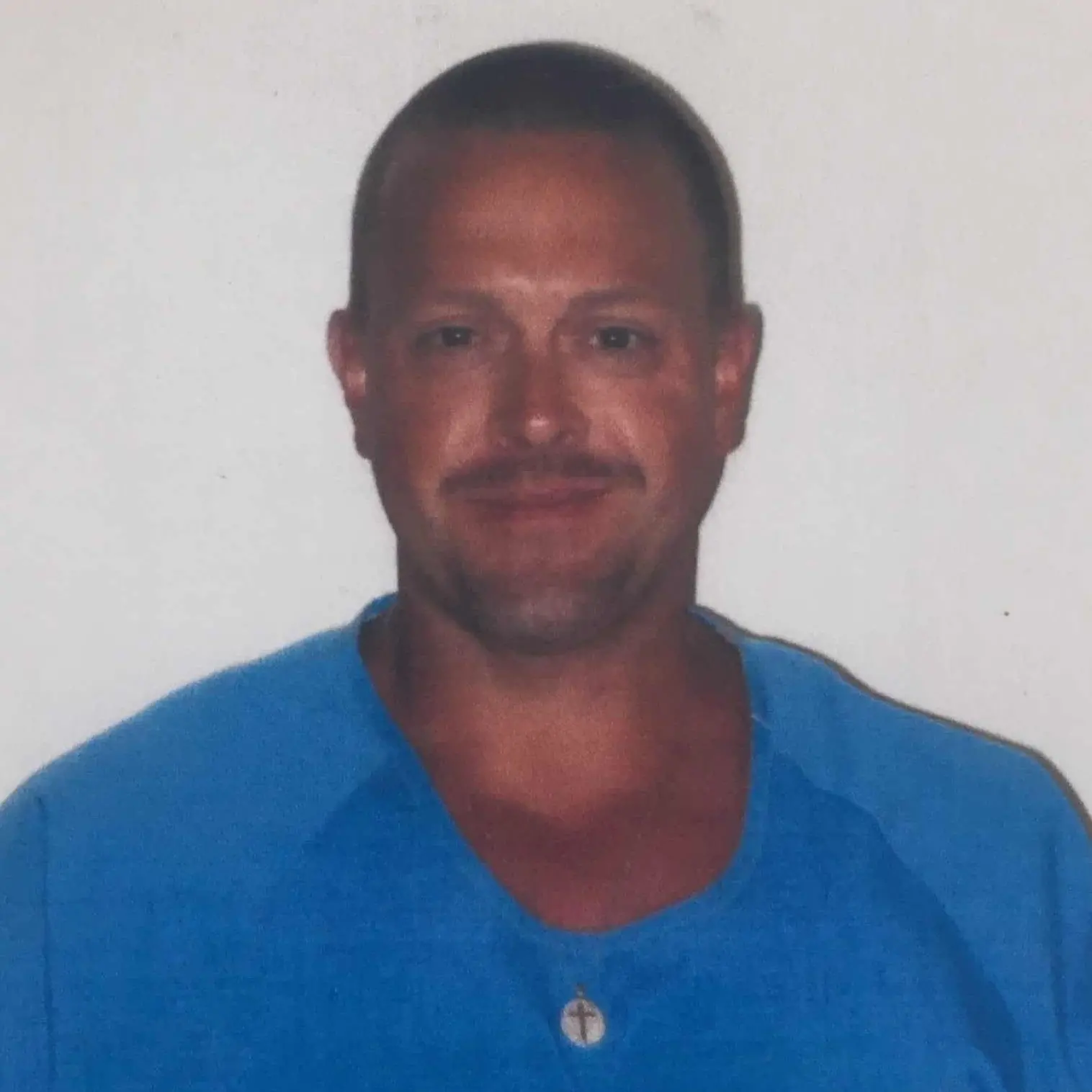 A man with short hair and a mustache, wearing a blue shirt and a necklace with a round pendant, stands in front of a plain white background, smiling slightly at the camera.
