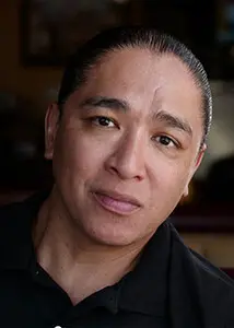 A man with dark hair pulled back, wearing a black shirt, looks directly at the camera with a neutral expression against a blurred indoor background.