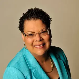 A smiling woman with short curly hair, wearing glasses, a turquoise blazer, a gold necklace, and earrings, poses against a plain beige background.