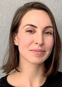 A woman with straight brown hair, parted to the side and cut to chin length, smiles gently. She is wearing a black top and is standing in front of a light grey textured wall.