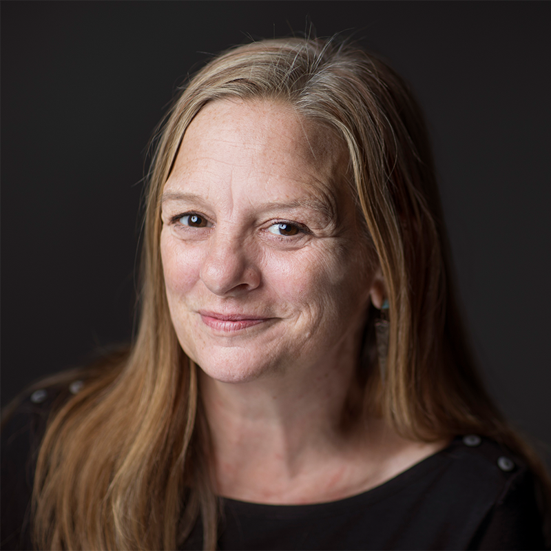 A middle-aged woman with long, light brown hair smiles gently at the camera. She is wearing a black top and is posed against a plain, dark background.