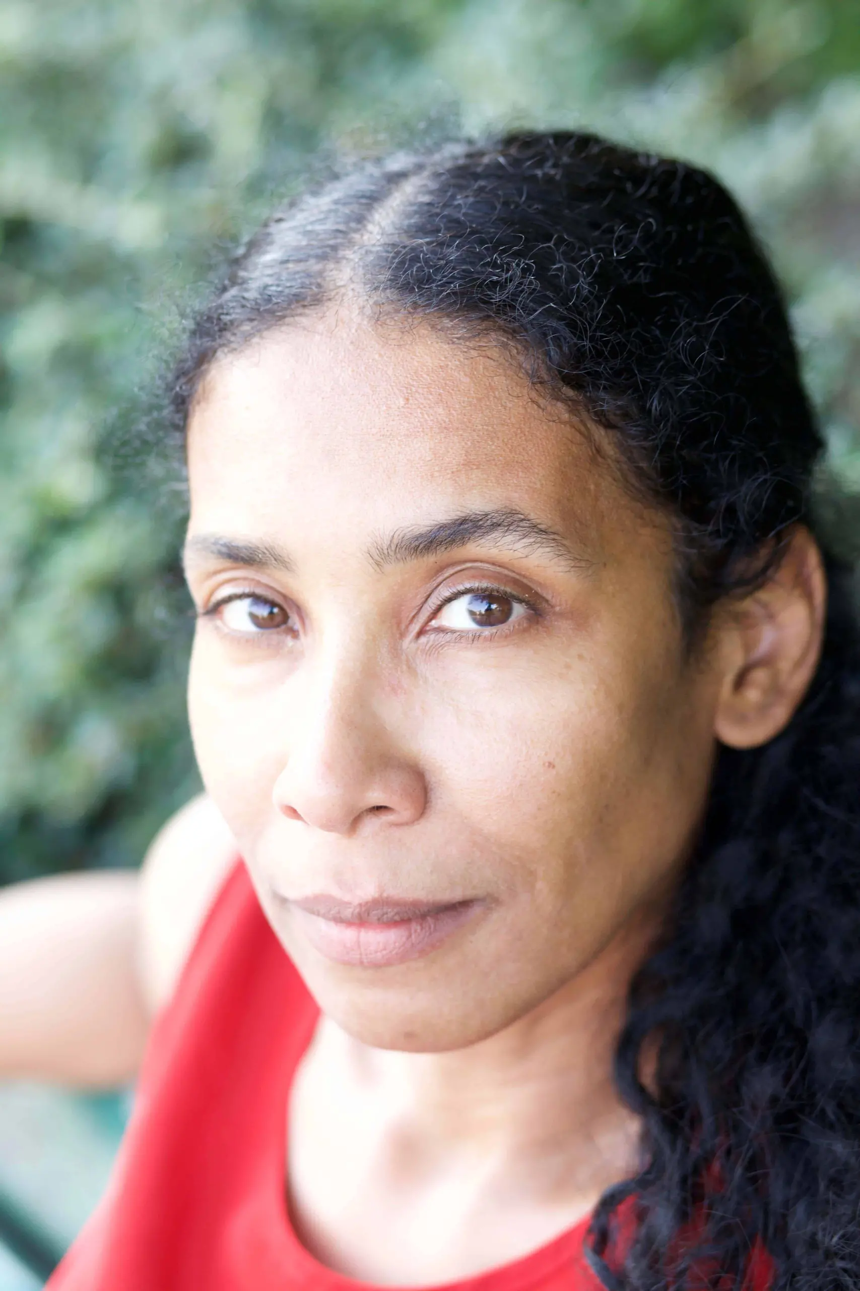A woman with long curly black hair and brown eyes gazes at the camera with a neutral expression. She is wearing a red top, and greenery is blurred in the background.