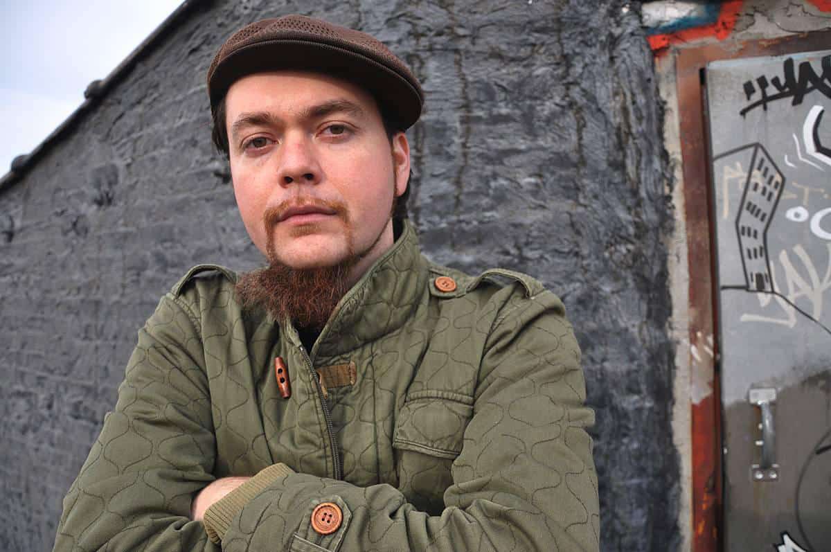 A man in a green quilted jacket and brown flat cap stands with arms crossed in front of a gray graffiti-covered wall, looking confidently at the camera.