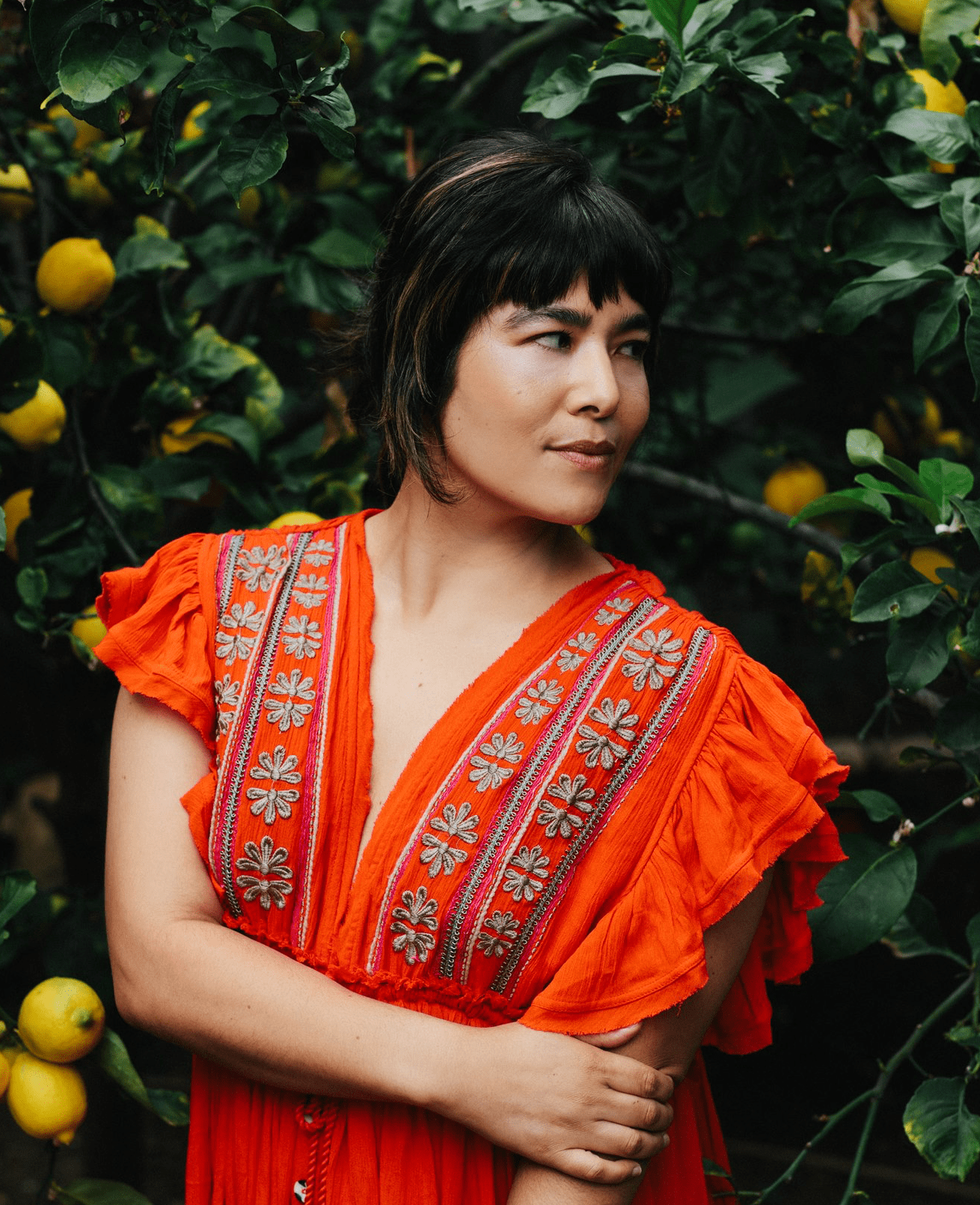 A person in a vibrant orange dress with embroidered patterns stands among lush green foliage and yellow lemons, looking thoughtfully to the side.