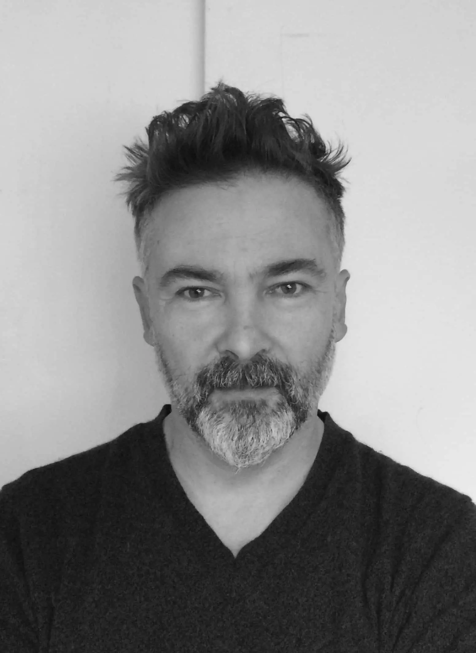 A man with short, dark, and slightly tousled hair and a full salt-and-pepper beard, wearing a dark V-neck shirt, stands against a plain light-colored background, looking directly at the camera.