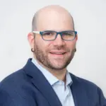 A man with a bald head, brown beard, and blue glasses, wearing a blue blazer and light-colored shirt, smiles at the camera against a plain light background, reflecting on the impact of educational censorship.
