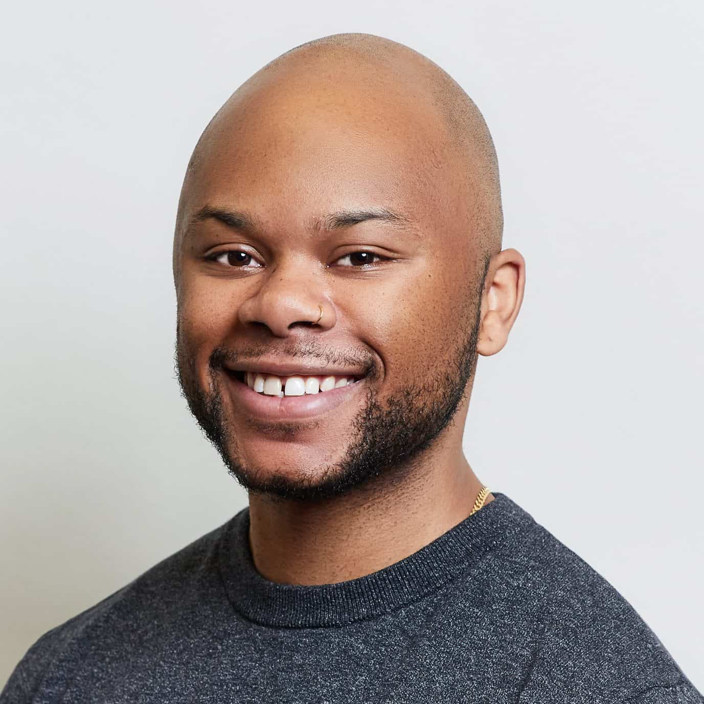 A smiling person with a shaved head, trimmed beard, and nose ring, wearing a dark grey shirt, poses in front of a plain light background.