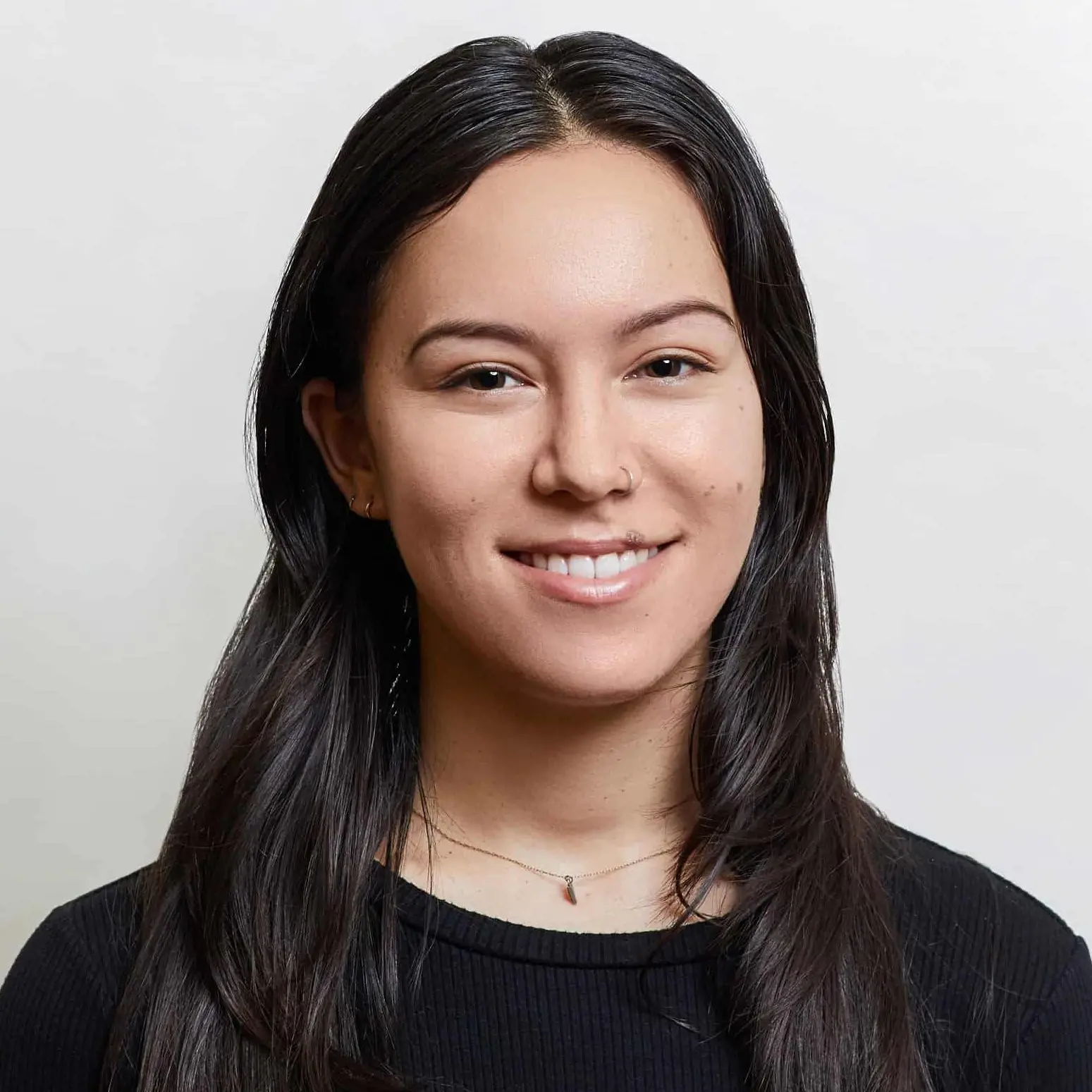 A woman with long dark hair and a light complexion smiles at the camera. She is wearing a black top and a delicate necklace, and is positioned against a plain, light-colored background.