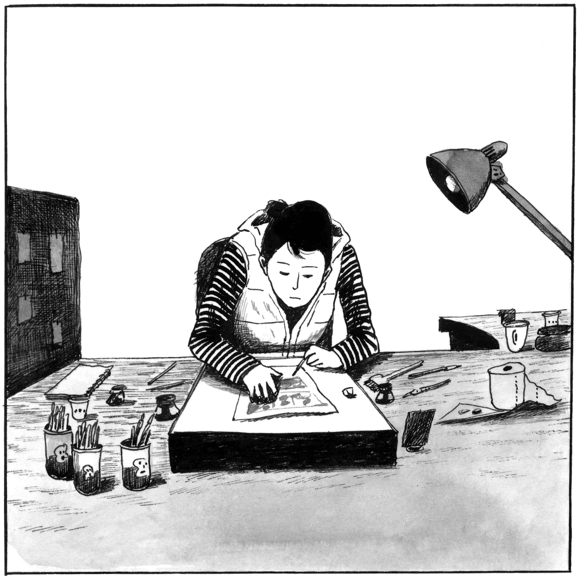 A person with short dark hair, wearing a striped shirt and vest, is intently drawing or working on a piece of art at a cluttered desk, illuminated by an angled desk lamp.