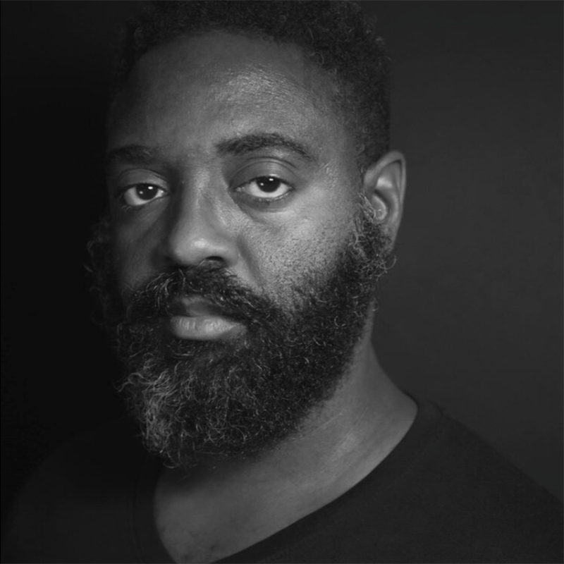 A black-and-white portrait of a man with short curly hair and a full, graying beard, wearing a dark V-neck shirt, looking directly at the camera against a dark background.