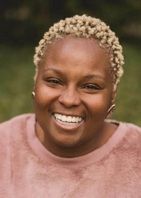 A person with short, curly blonde hair smiles warmly at the camera, wearing a pink fuzzy sweater, with a natural, blurred green background.