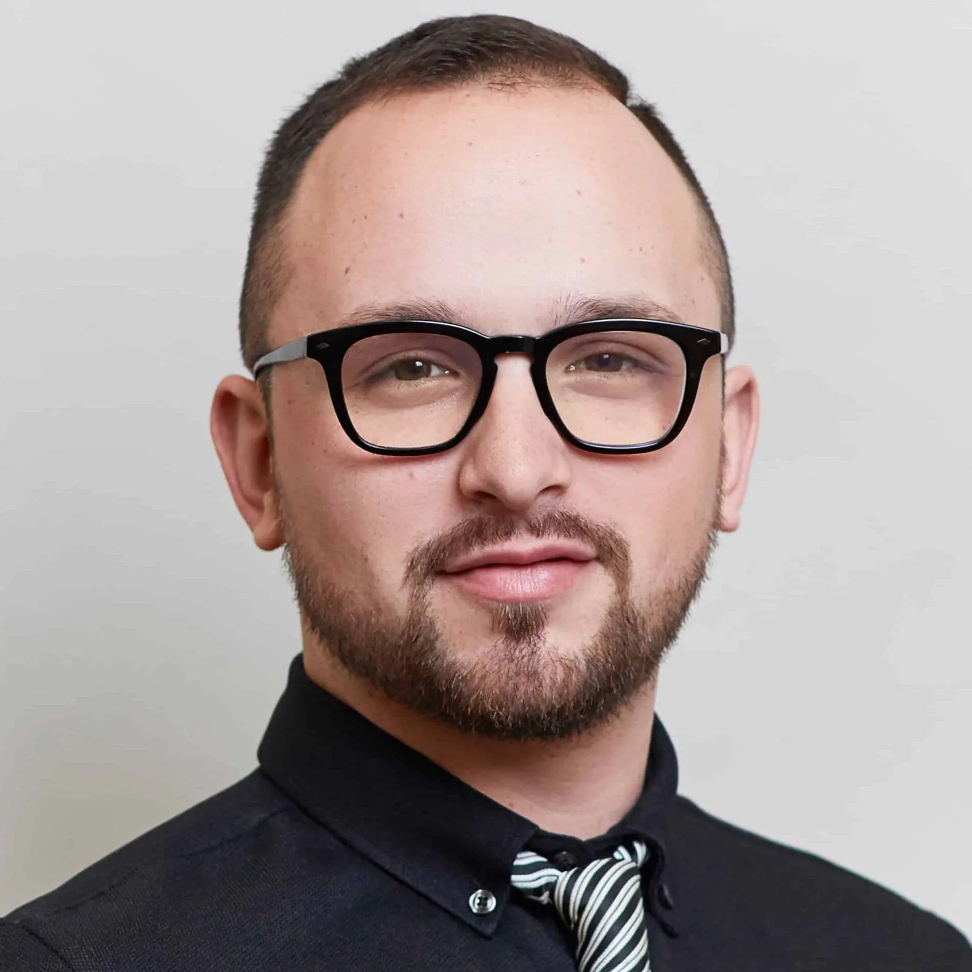 A person with short hair, a trimmed beard, and black eyeglasses—wearing a black shirt and striped tie—looks at the camera with a neutral expression, evoking the thoughtful energy of NYC Free Expression Advocacy Institute PEN.
