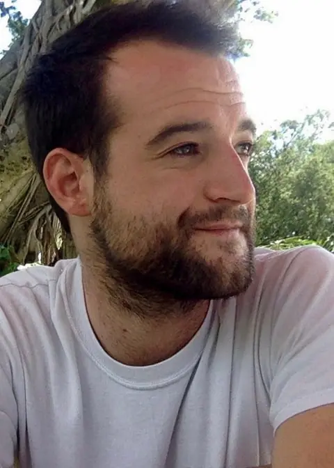 A man with short dark hair and a beard, wearing a white t-shirt, sits outdoors and looks to the side with a slight smile. Trees and greenery are visible in the background.