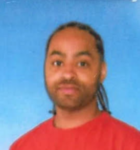 A person with braided hair, a beard, and mustache is wearing a red shirt and posing in front of a solid blue background, reflecting on prison justice amid the covid pandemic.