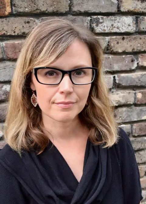 A woman with shoulder-length blonde hair, wearing black glasses, a black top, and dangling earrings, stands in front of a brick wall, looking at the camera with a neutral expression.