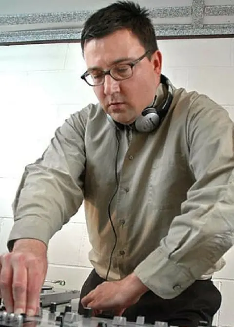 A man wearing glasses and headphones adjusts controls on a DJ mixer. He is dressed in a light-colored button-up shirt and is focused on his work, standing in front of a white brick wall.