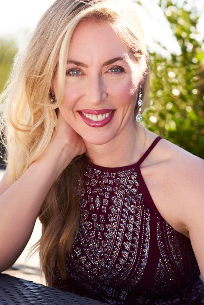 A smiling woman with long blonde hair wears a sleeveless, sparkling burgundy dress and dangling earrings, posing outdoors with sunlight highlighting her face—Alethea Black—against a blurred green background.
