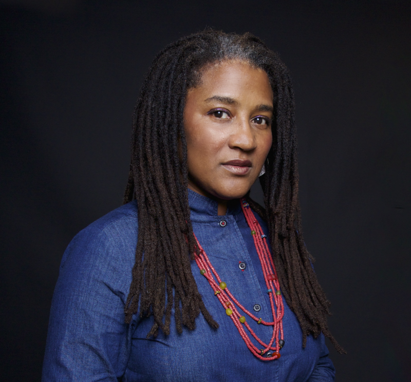 A woman with long, dark, braided hair wears a blue button-up top and layered red beaded necklaces, posing against a dark background with a neutral expression.