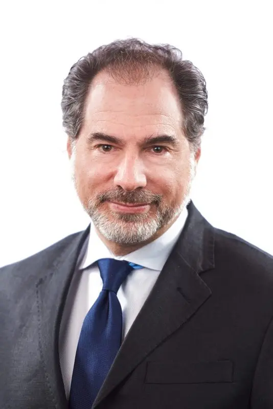 A middle-aged man with dark hair and a beard, wearing a dark suit, white shirt, and blue tie, is smiling slightly against a plain white background.