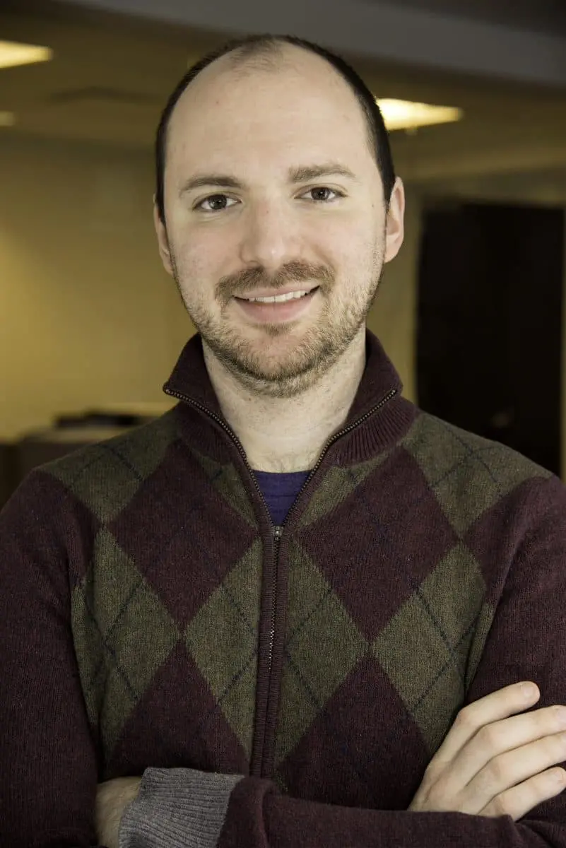 A man with a receding hairline and short beard is smiling with his arms crossed, wearing a maroon and olive argyle sweater in an indoor setting with soft lighting.