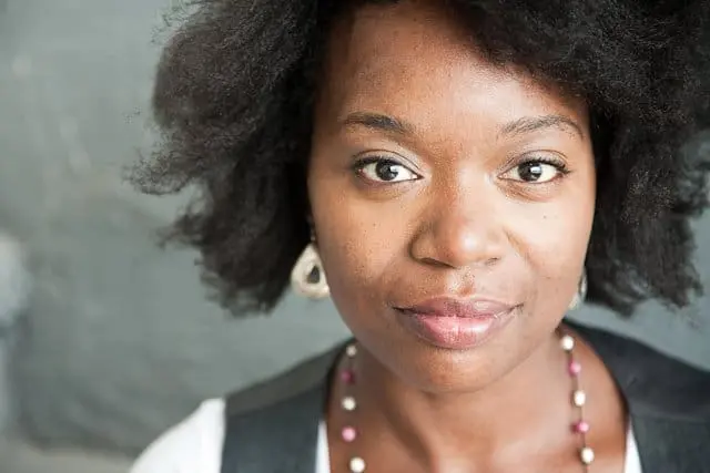 A woman with natural, curly black hair and brown eyes looks at the camera, wearing a white top, a black vest, a beaded necklace, and round earrings, against a blurred gray background.