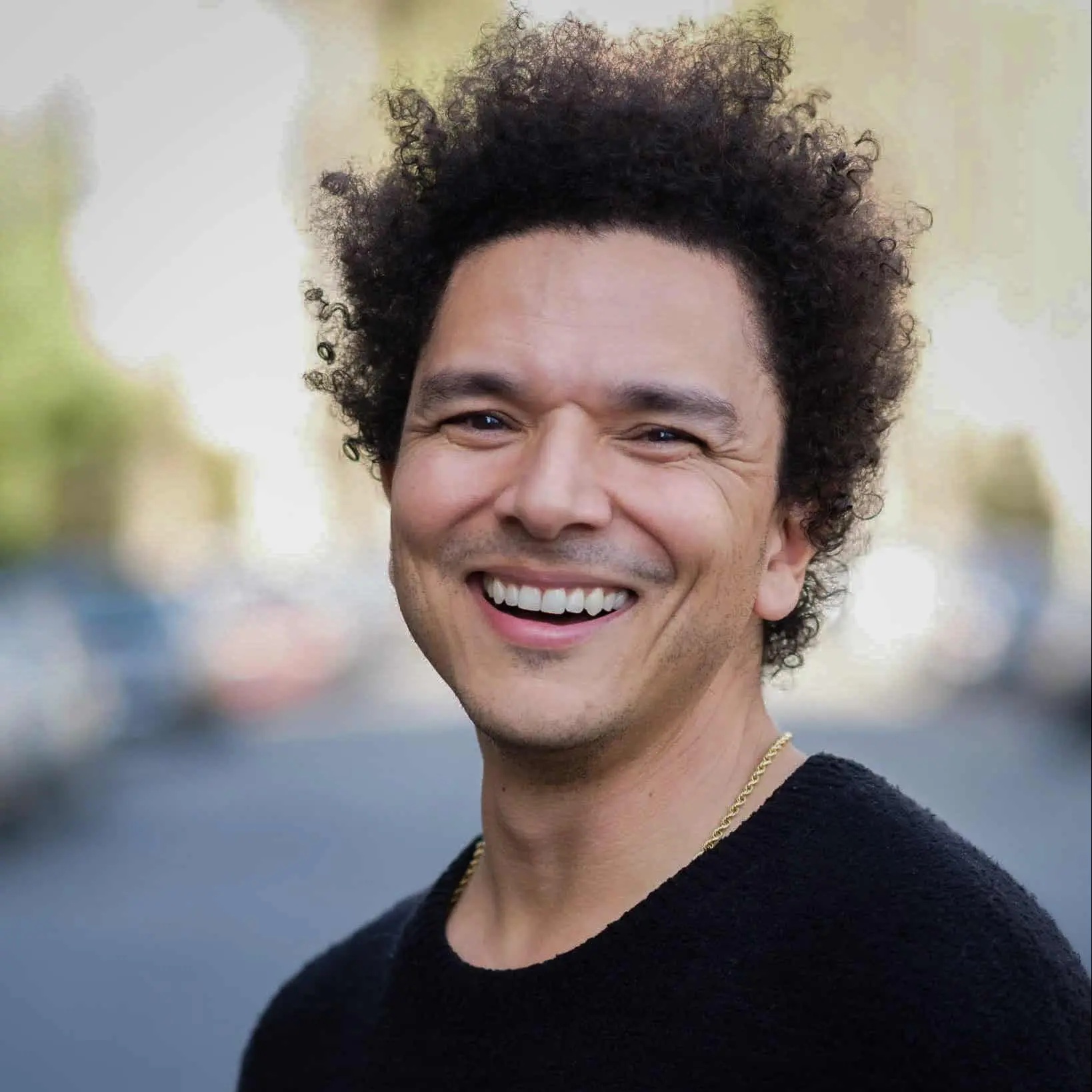 A man with curly hair and a black sweater smiles warmly while standing outdoors, with a blurred street and trees in the background.
