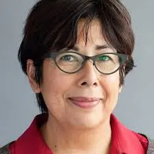 A middle-aged person with short dark hair, wearing black-rimmed glasses and a red collared shirt, smiles gently in front of a plain gray background.