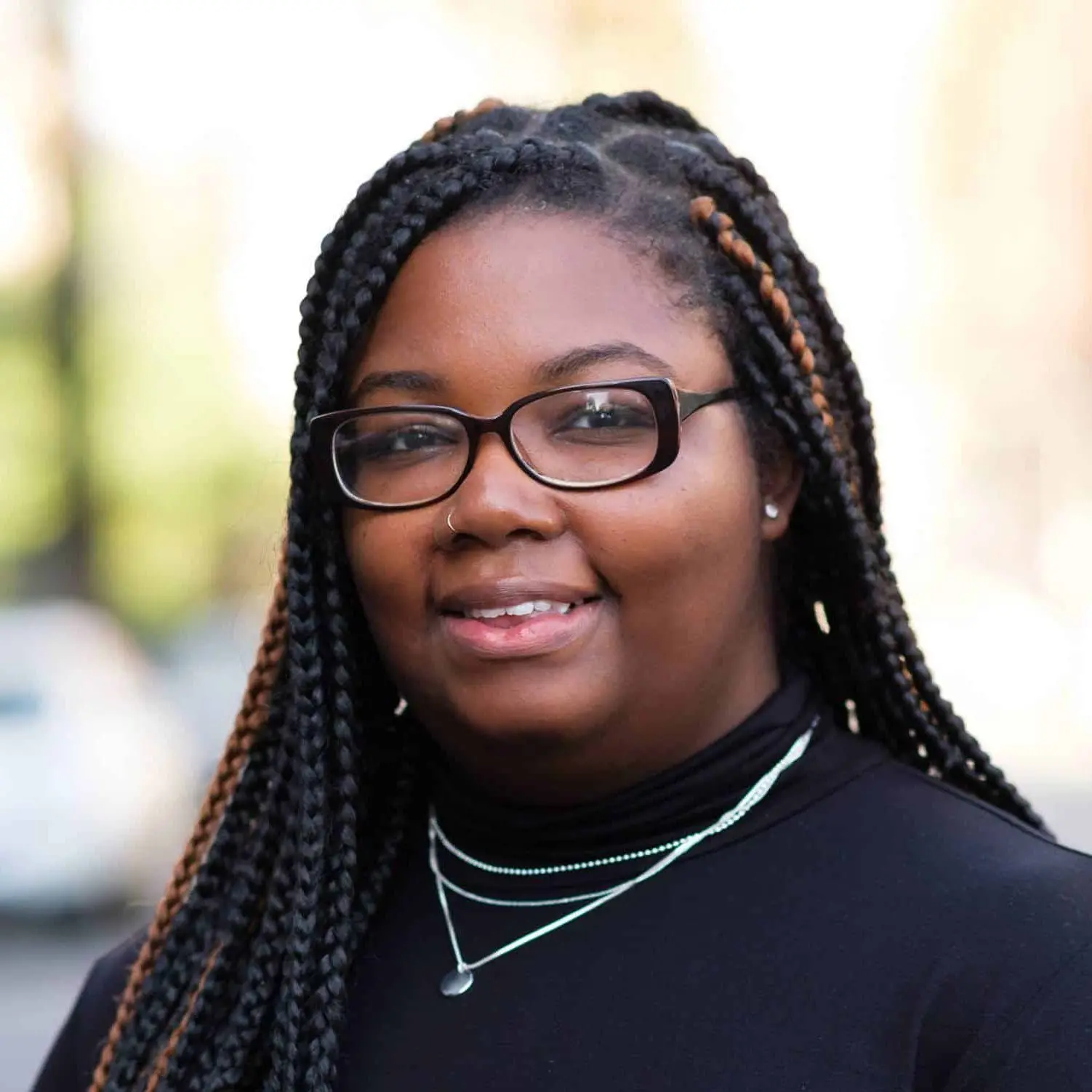 A person with long, braided hair, wearing glasses, a black top, and layered necklaces, smiles outdoors with a blurred background of trees and buildings.