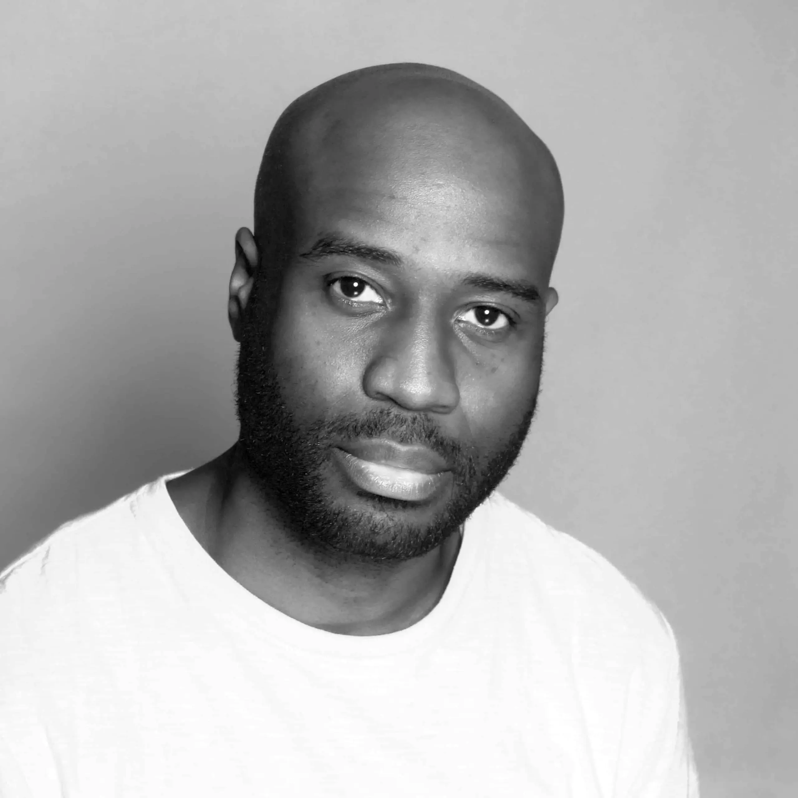 A man with a beard and shaved head wearing a light-colored T-shirt looks toward the camera against a plain background. The image is in black and white.