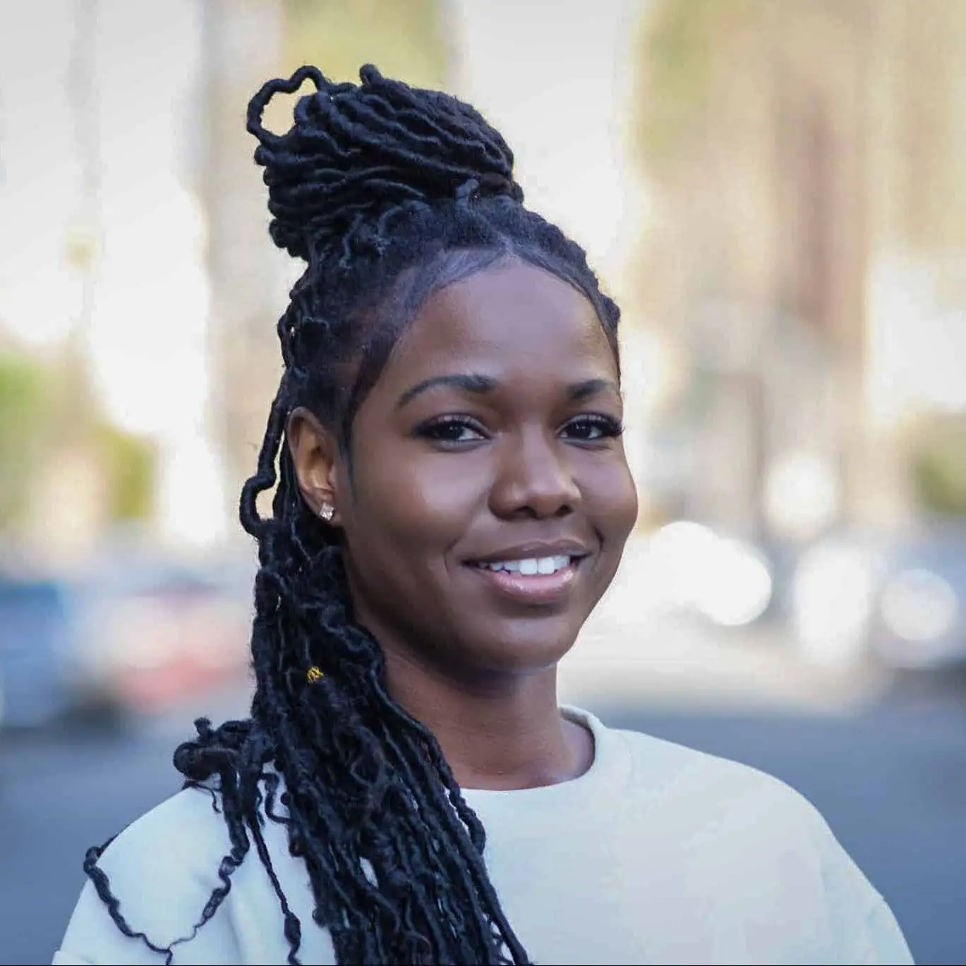 A woman with long, styled black locs and a white top smiles outdoors, with a blurred background of trees and parked cars.