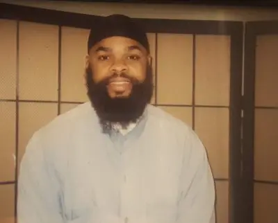 A man with a full beard, wearing a dark head covering and a light-colored shirt, sits in front of a folding room divider with a grid pattern, reflecting on voting in prison during the covid-19 pandemic.