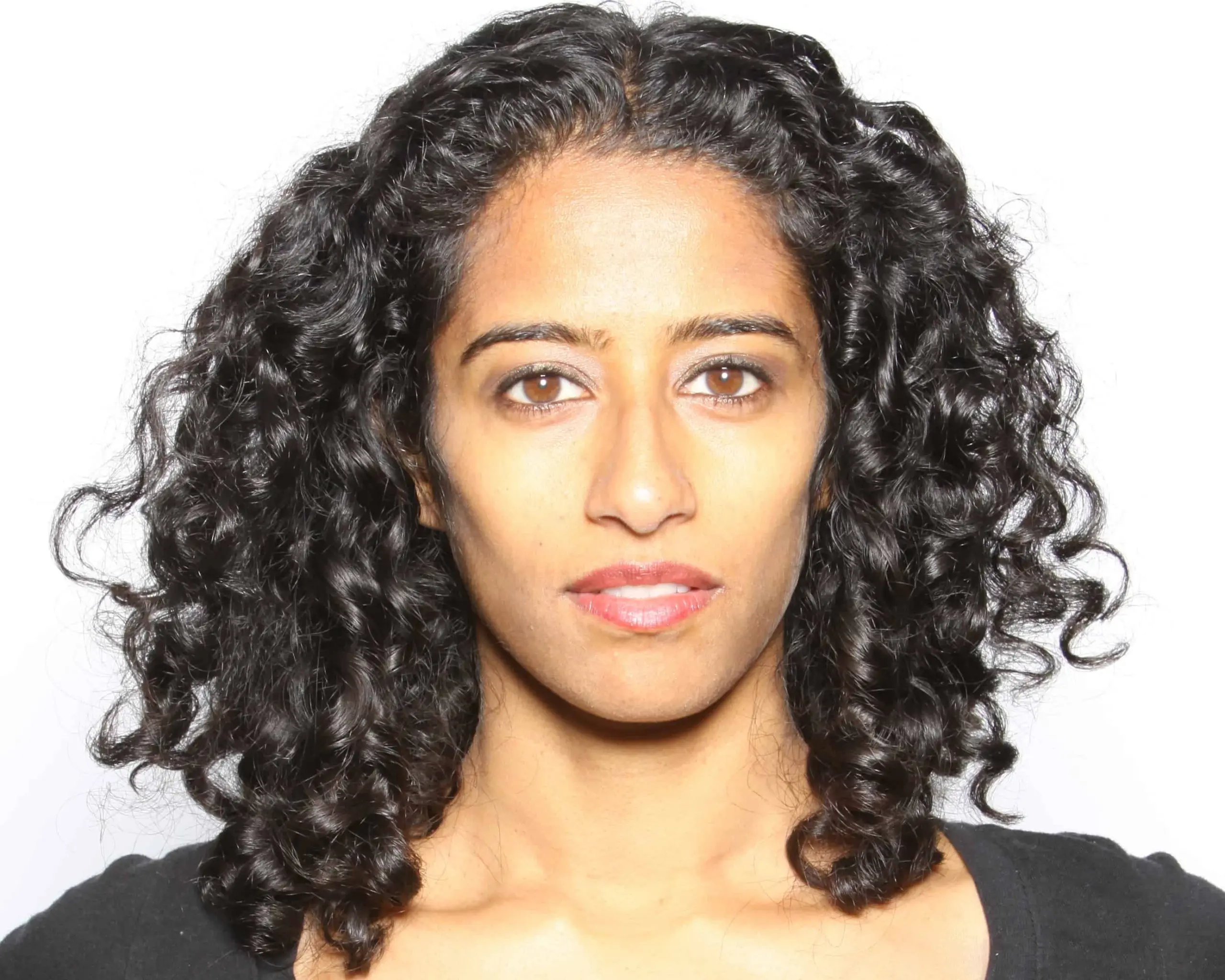 A woman with medium skin tone and curly dark hair looks directly at the camera with a neutral expression, standing against a plain white background. She is wearing a black top.